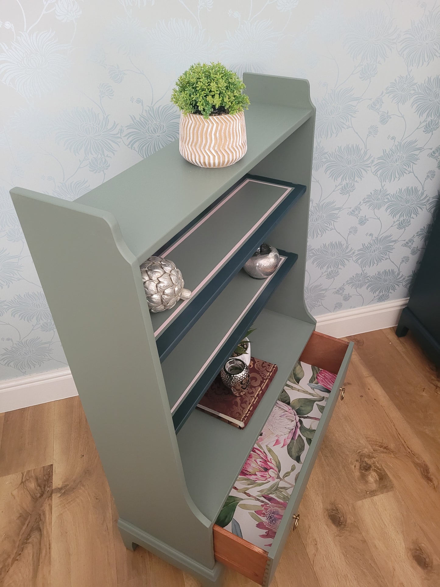 Beautifully upcycled green bookshelf with floral wallpaper backing. The shelves display decorative items and the bottom drawer features elegant gold ring pulls. Perfect for adding a touch of nature-inspired charm to any living room, bedroom, or hallway.