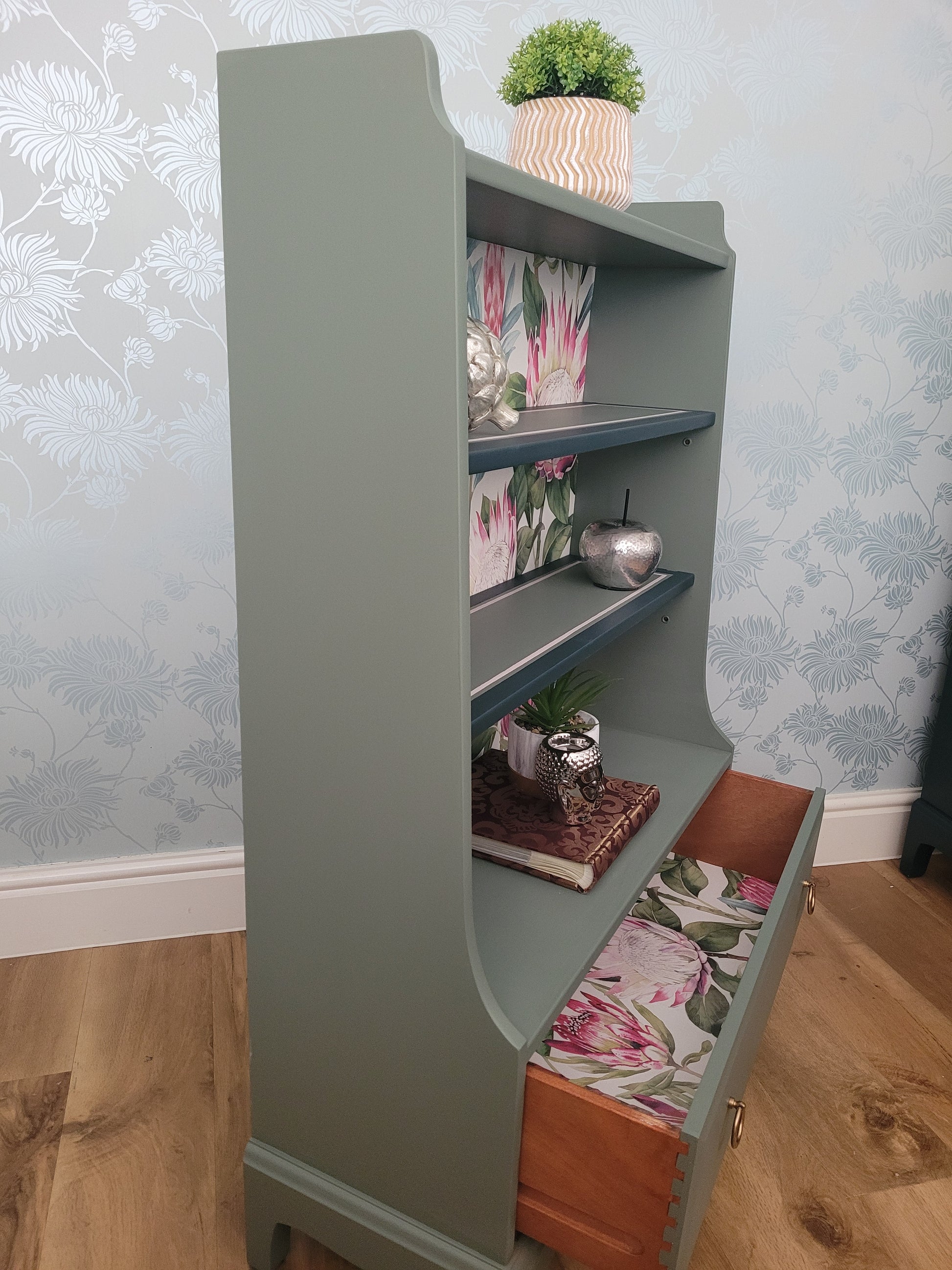 Beautifully upcycled green bookshelf with floral wallpaper backing. The shelves display decorative items and the bottom drawer features elegant gold ring pulls. Perfect for adding a touch of nature-inspired charm to any living room, bedroom, or hallway.