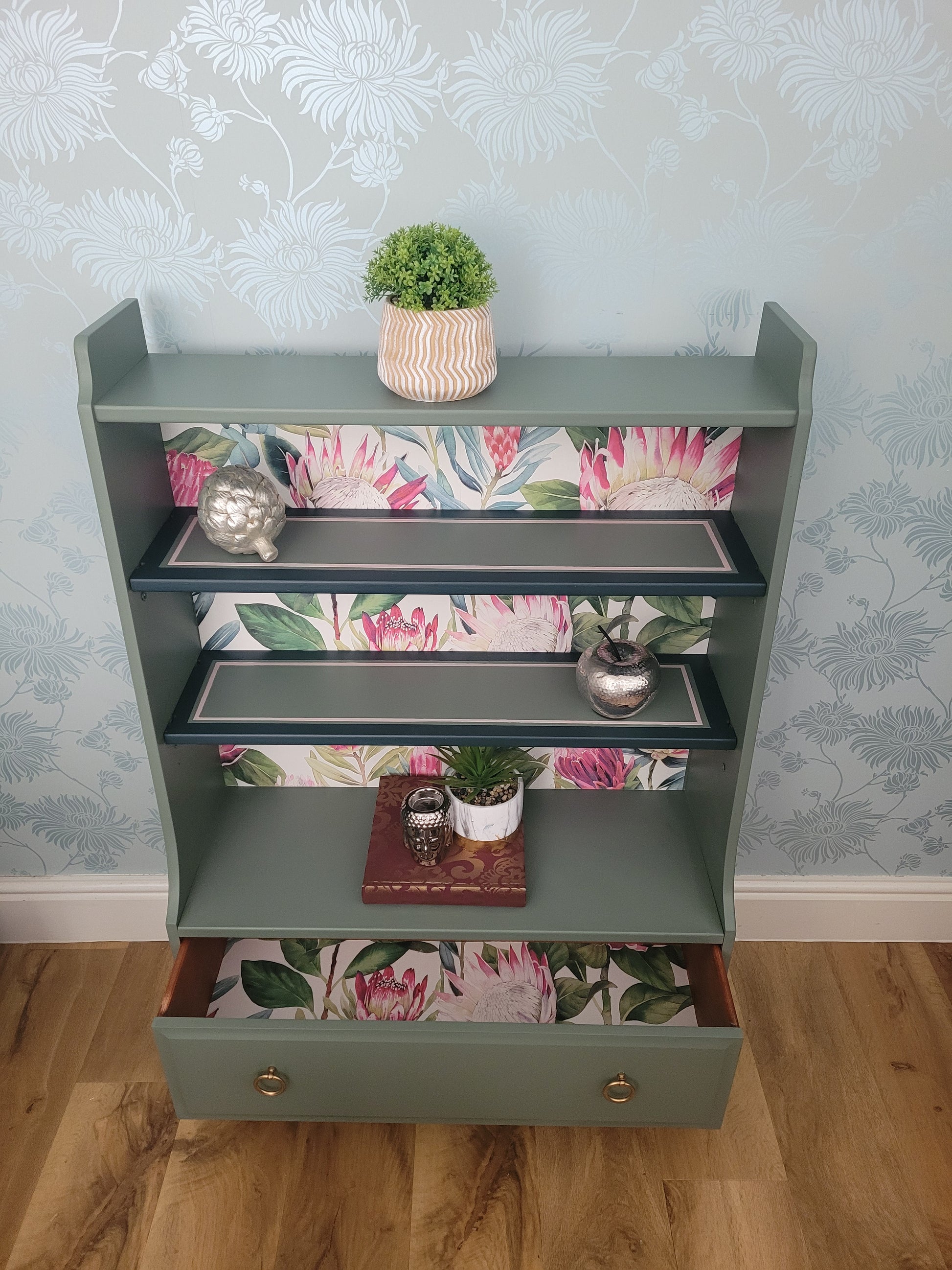 Beautifully upcycled green bookshelf with floral wallpaper backing. The shelves display decorative items and the bottom drawer features elegant gold ring pulls. Perfect for adding a touch of nature-inspired charm to any living room, bedroom, or hallway.