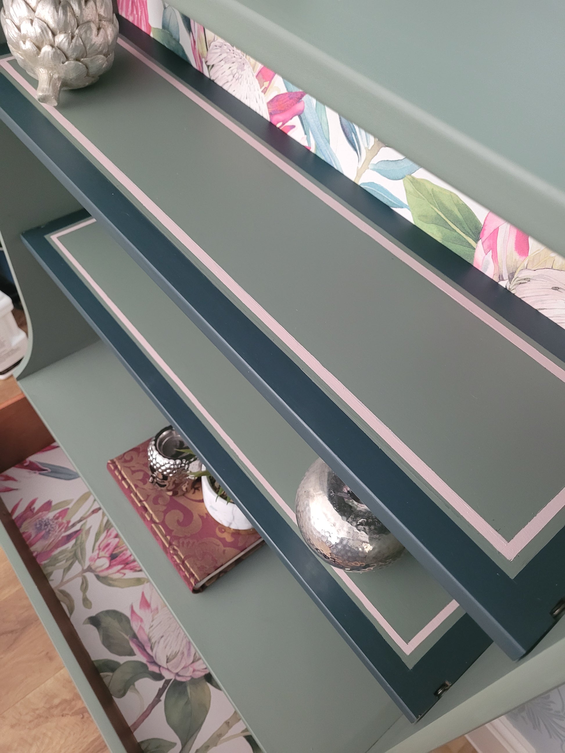 Beautifully upcycled green bookshelf with floral wallpaper backing. The shelves display decorative items and the bottom drawer features elegant gold ring pulls. Perfect for adding a touch of nature-inspired charm to any living room, bedroom, or hallway.