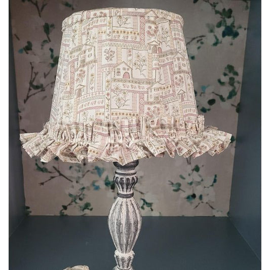 Tapered hand-sewn fabric French drum lampshade featuring alpine flowers and animals in a whimsical design. Finished with ruffle trim for a charming touch, adding a unique and cosy element to any table lamp. Crafted with Liberty London fabric.