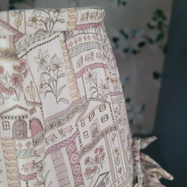 Tapered hand-sewn fabric French drum lampshade featuring alpine flowers and animals in a whimsical design. Finished with ruffle trim for a charming touch, adding a unique and cosy element to any table lamp. Crafted with Liberty London fabric.