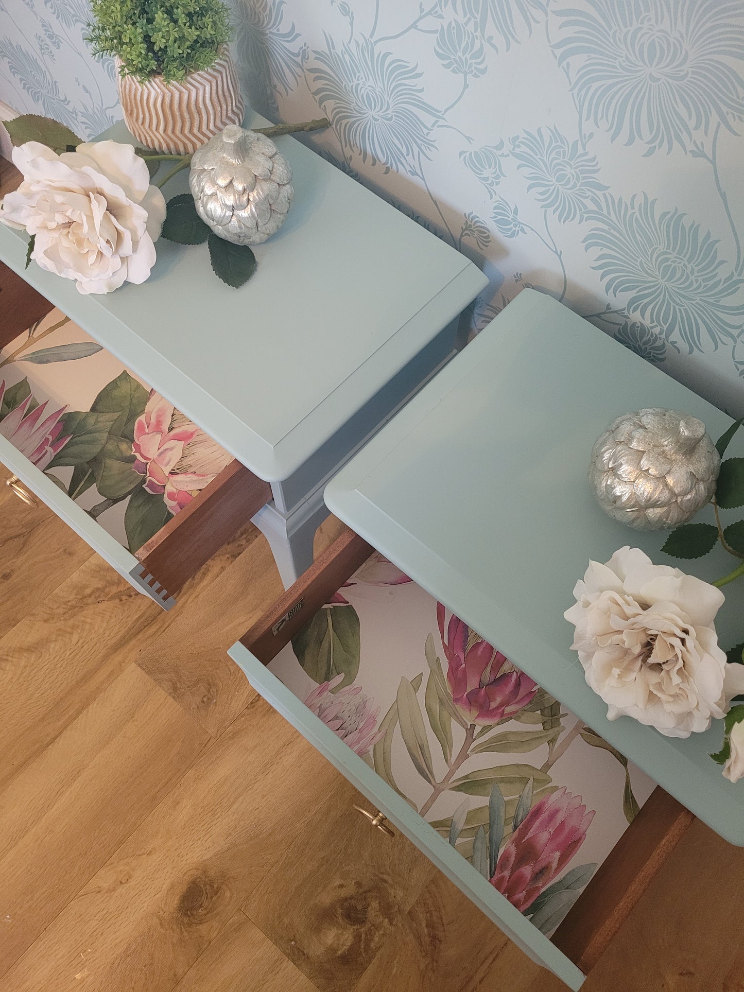 A top-down view of a light blue painted wooden chest of drawers with floral wallpaper and decorative flowers.