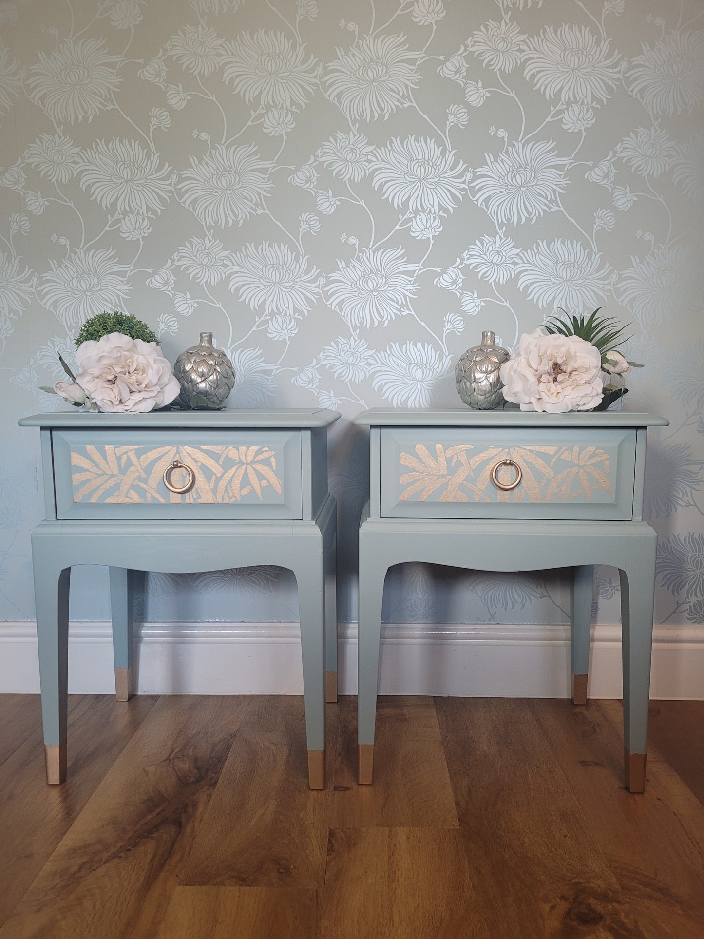 A pair of matching light blue bedside tables with single drawers, decorated with floral wallpaper and faux flowers, shown on a wooden floor.