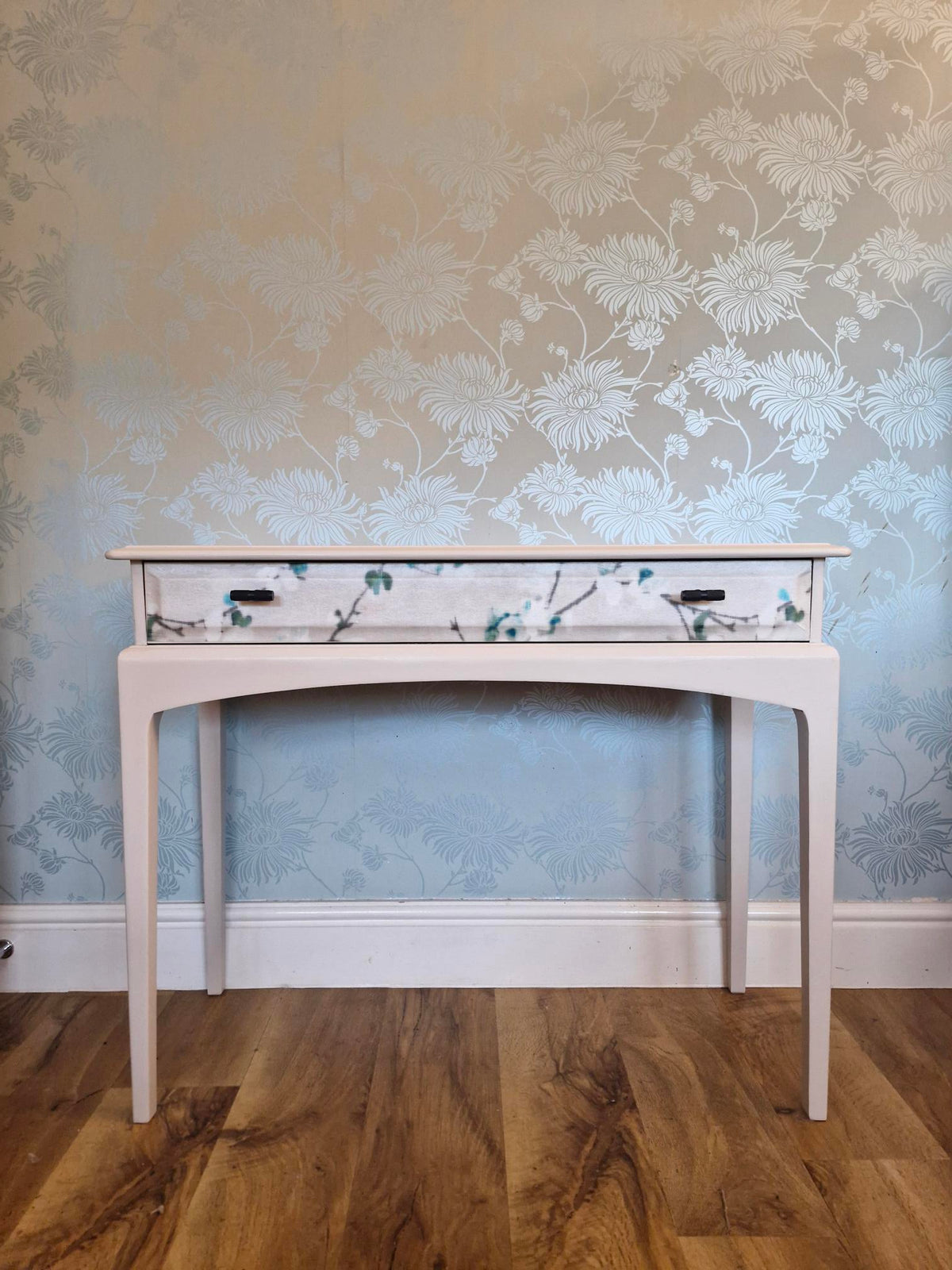 A cream console table with a floral-patterned drawer front, shown from a top-down angle on a wood floor against patterned wallpaper.