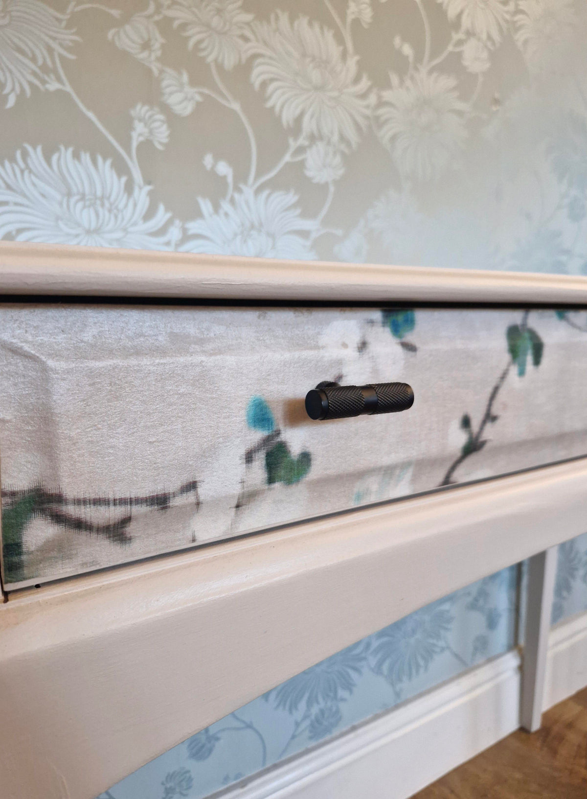 Close-up of a cream-colored wooden furniture piece, likely a dresser or cabinet drawer, featuring a floral-patterned fabric drawer front with a small black metal pull handle.
