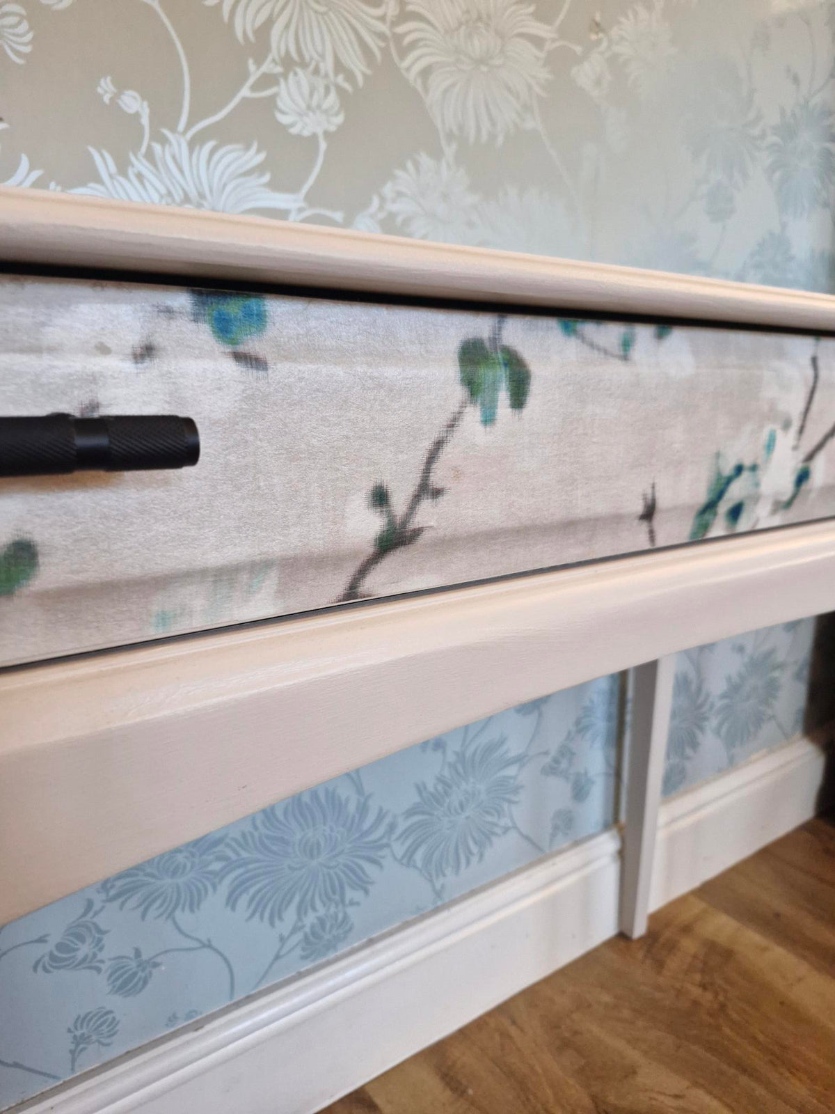 A close-up view of a console table drawer front trim, showing  floral-patterned decoupaged wallpaper. A black handle is visible at the bottom, and the surrounding area includes patterned wallpaper and a wood floor.