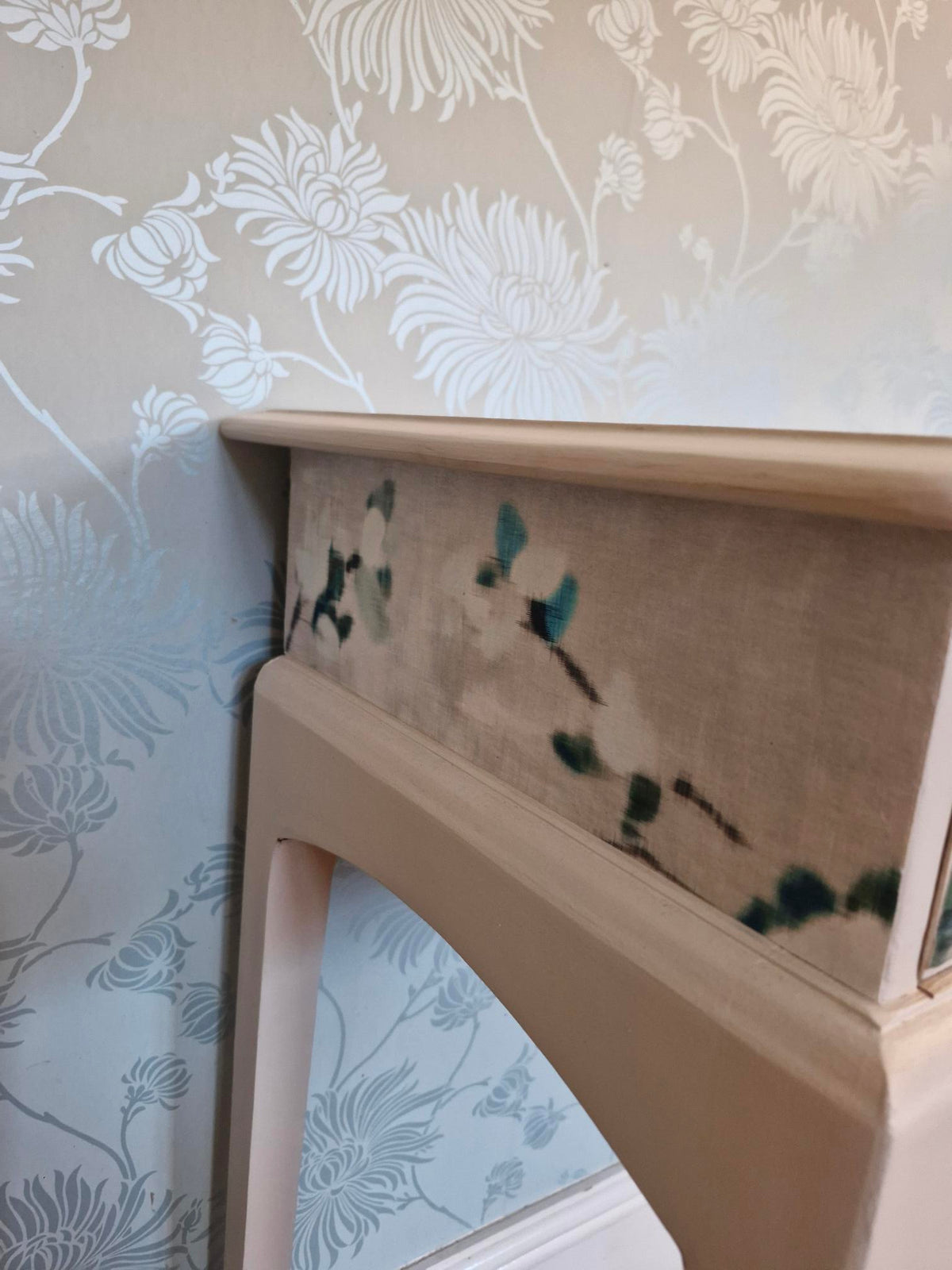 Close-up of a light wood furniture leg or frame corner resting against a cream floral wallpapered wall and floor, with a patterned fabric panel visible between the legs.