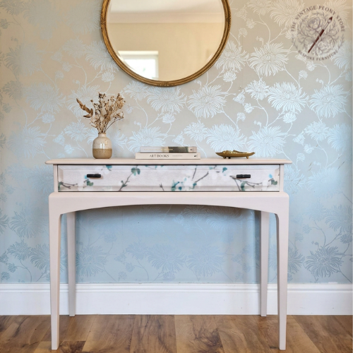A cream console table with a floral-patterned drawer front and curved legs, styled in a room with light blue patterned wallpaper, a round gold-framed mirror, and a wooden floor.