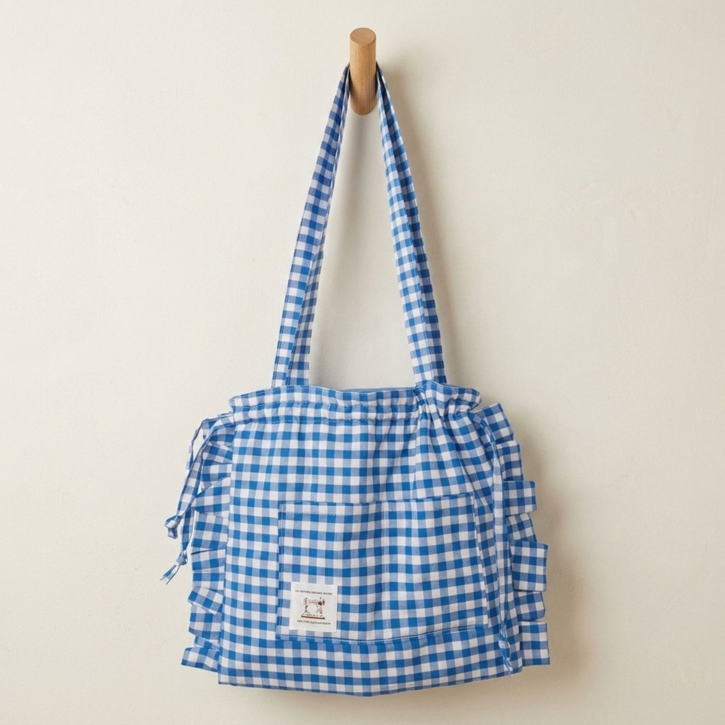 Blue gingham tote bag set against a plain cream wall on a wooden hook.