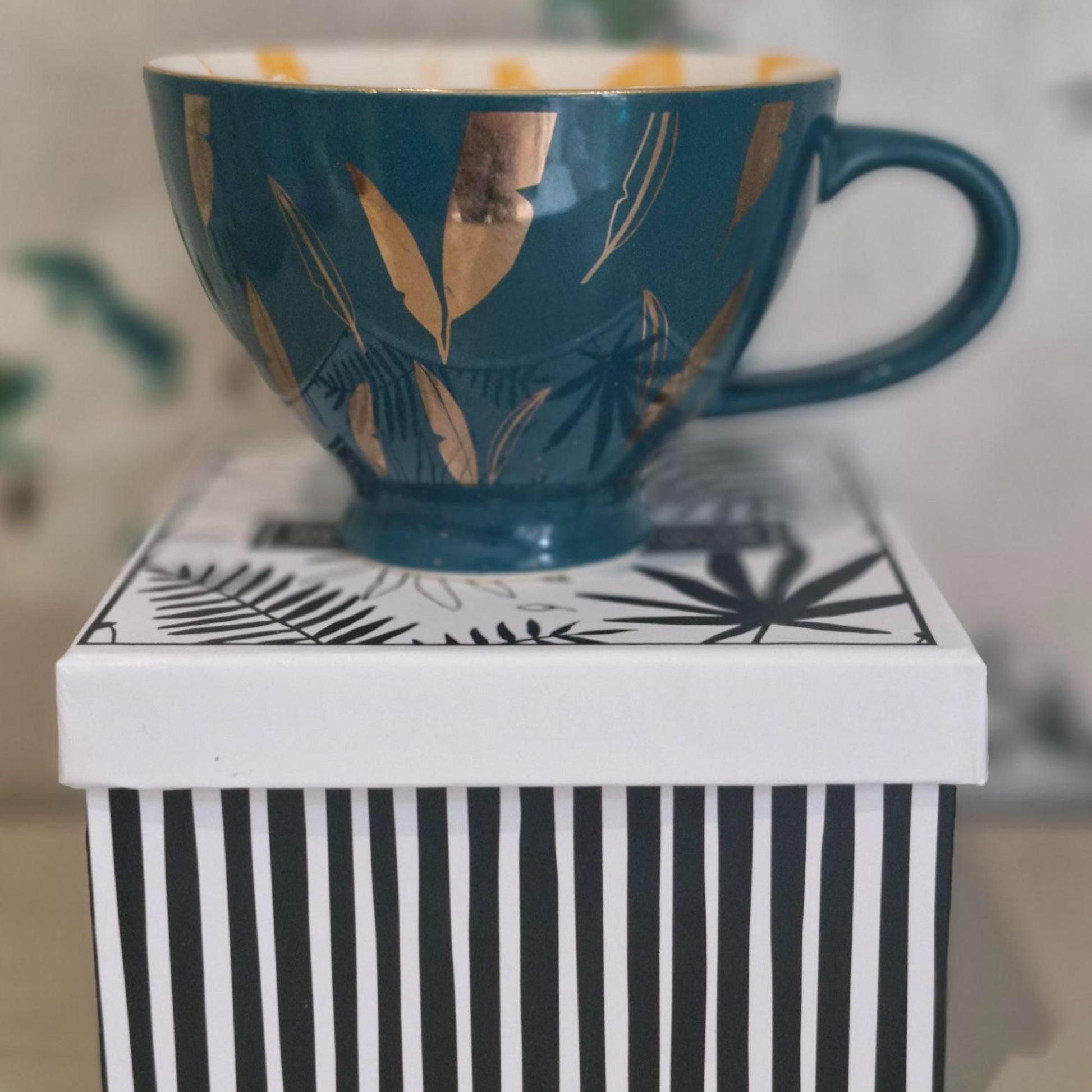 Green / Blue mug with gold leaf design on a white surface with black stripes