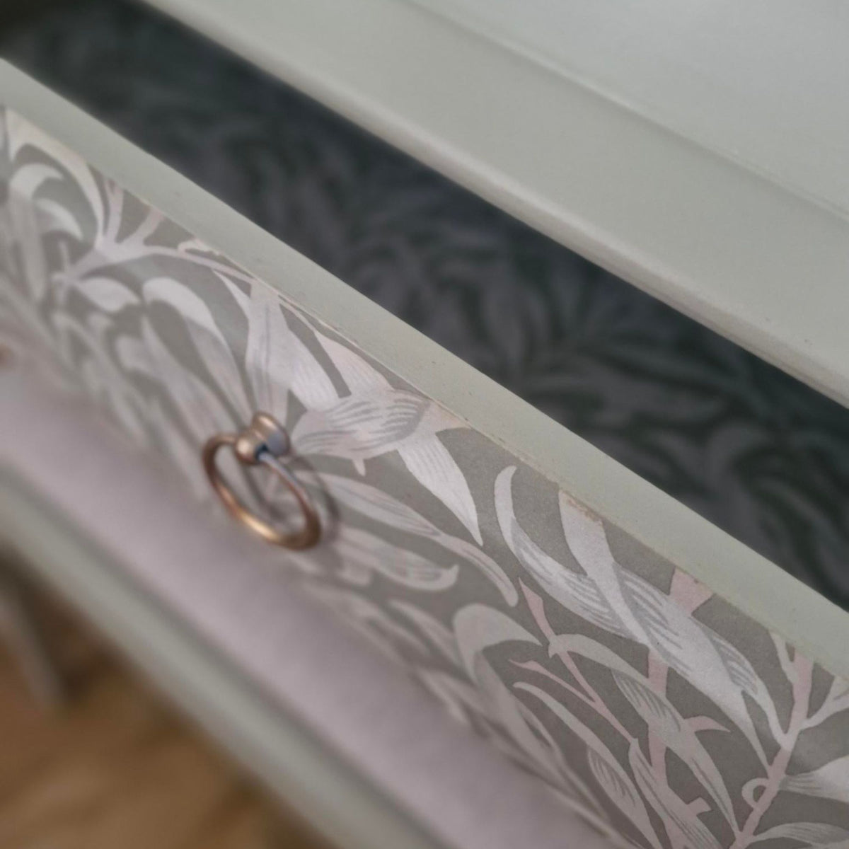 Close-up of a drawer front with a green and white botanical leaf pattern, a light gray frame, and a round metallic ring pull.