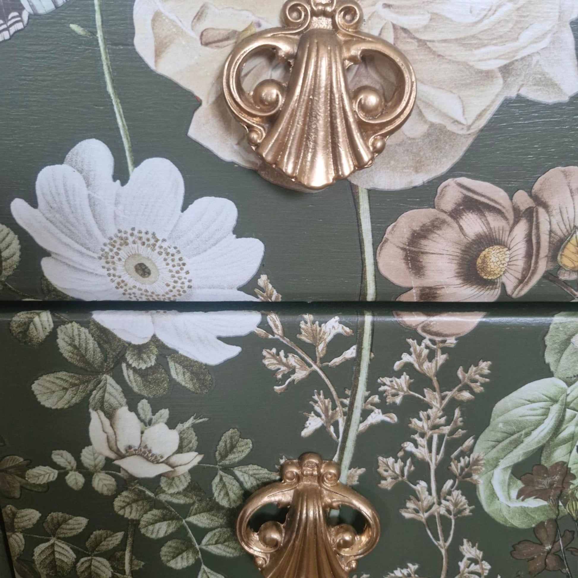 Close-up of a decorative floral surface with large white and muted pink flowers on a dark green background, featuring ornate metallic gold shell-shaped hardware.