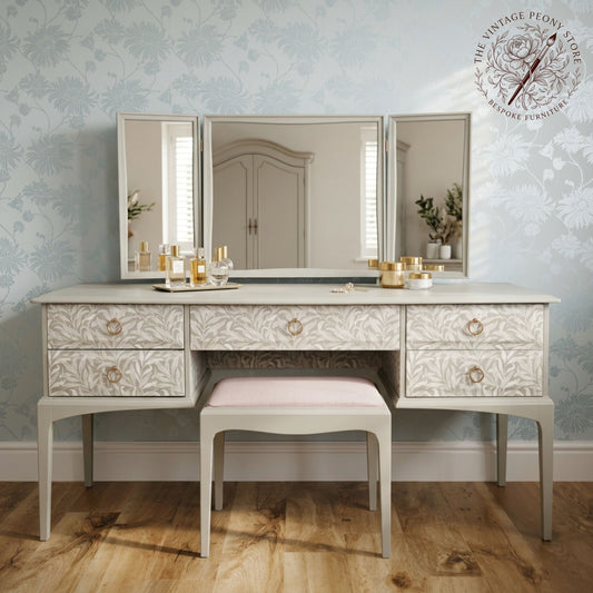 A vintage-style vanity table with a matching stool, featuring a three-panel mirror and ornate floral engraving on a light gray wood. The vanity is styled with cosmetic bottles and jars on a light wood floor against a blue floral wallpaper background.