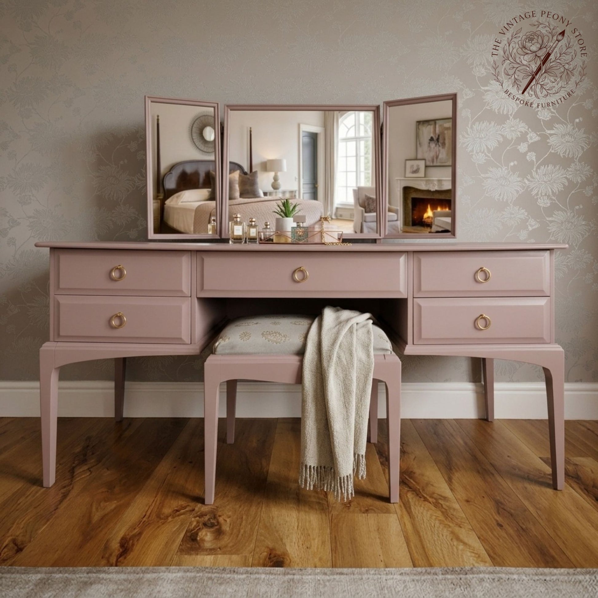 A dusty pink wooden vanity table with three mirrors and a matching stool, styled in a vintage bedroom setting with a wooden floor and patterned wallpaper.