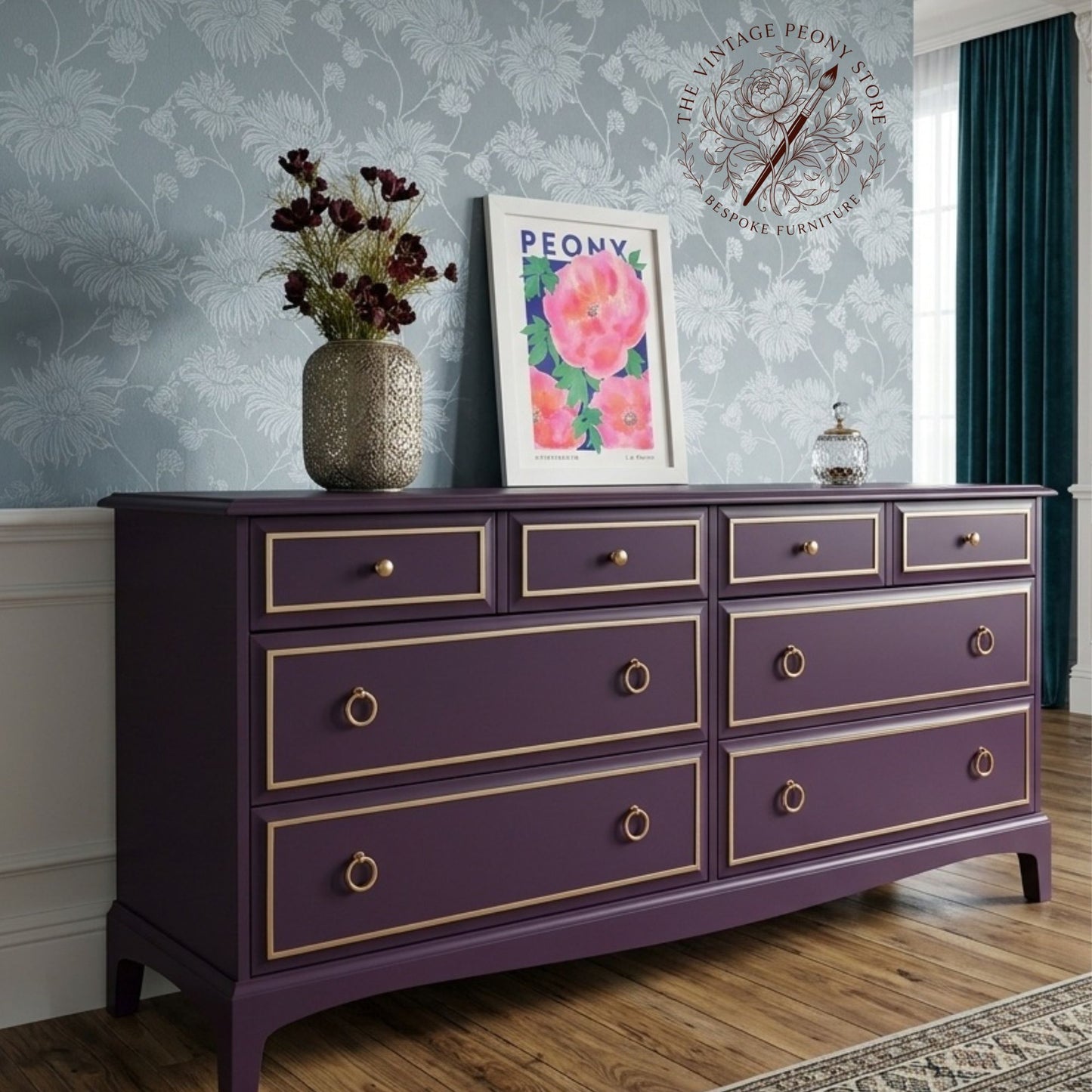 A purple six-drawer dresser with gold trim and brass ring pulls, styled in a room with floral wallpaper, a vase of flowers, and a framed print.