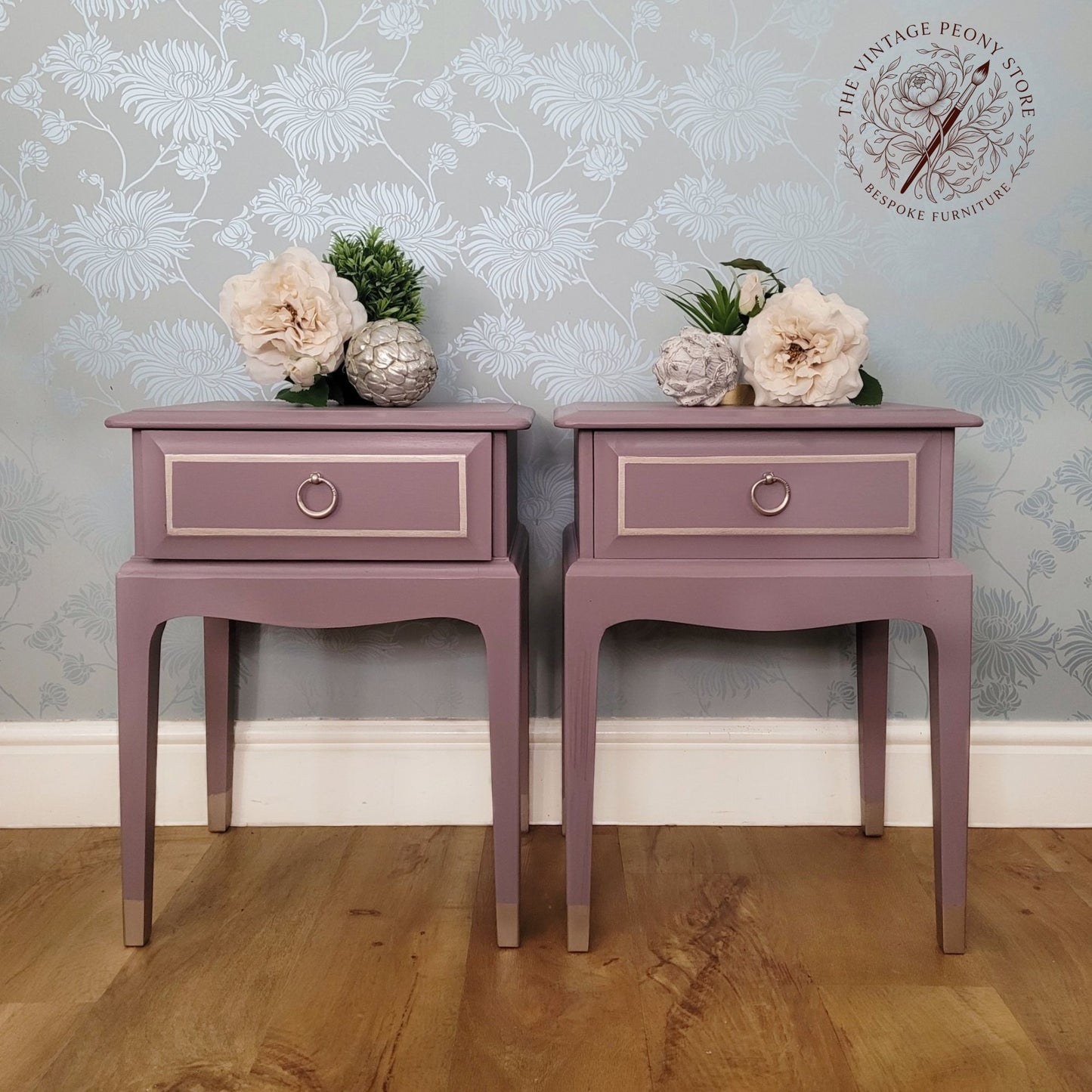A pair of matching dusty mauve side tables with single drawers and circular metal knobs, styled with flowers on top against a floral wallpaper background. A watermark logo for 'The Vintage Peony Store' is visible in the top right corner.