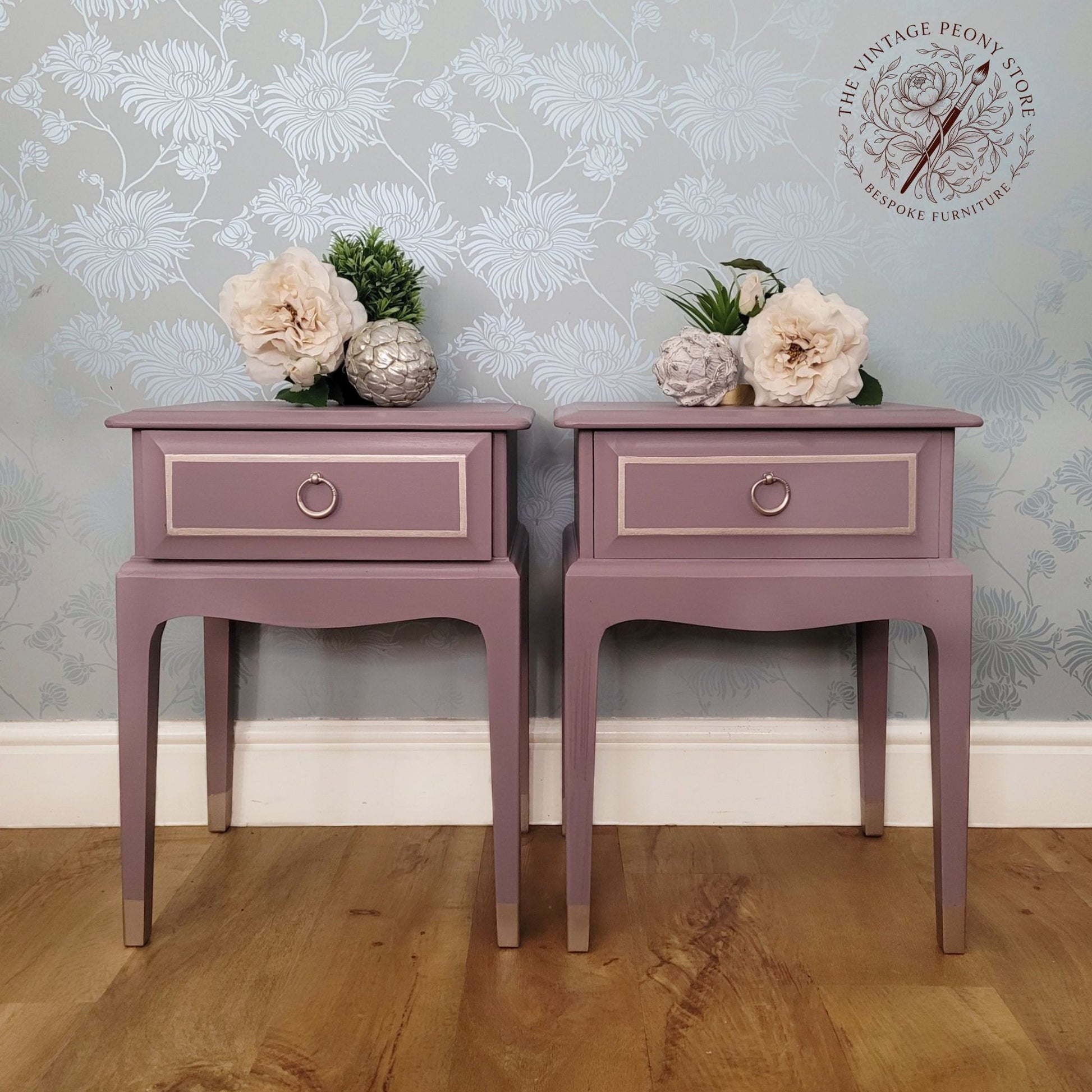 A pair of matching dusty mauve side tables with single drawers and circular metal knobs, styled with flowers on top against a floral wallpaper background. A watermark logo for 'The Vintage Peony Store' is visible in the top right corner.