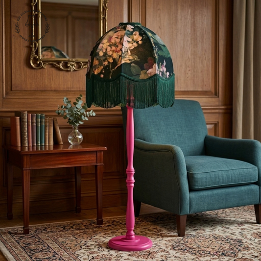 A vintage-style floor lamp with a dark green fringed shade featuring a floral print and a glossy pink turned wood stand, placed in a classic room setting next to a teal armchair and a wooden side table.