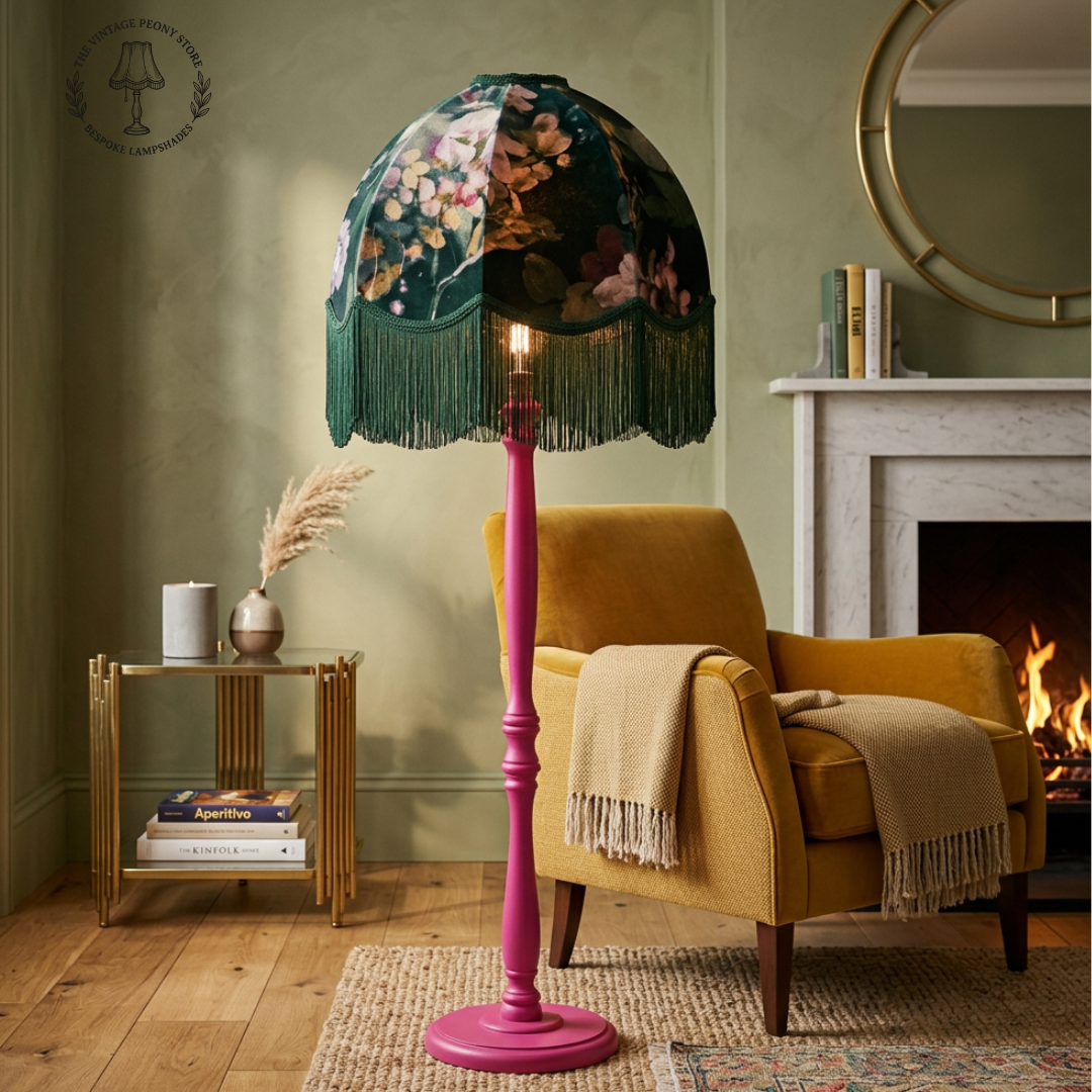 A vintage-style dome floor lamp with a dark green floral velvet shade and fringe, mounted on a bright pink pole, shown in a cozy living room next to a yellow armchair and fireplace.