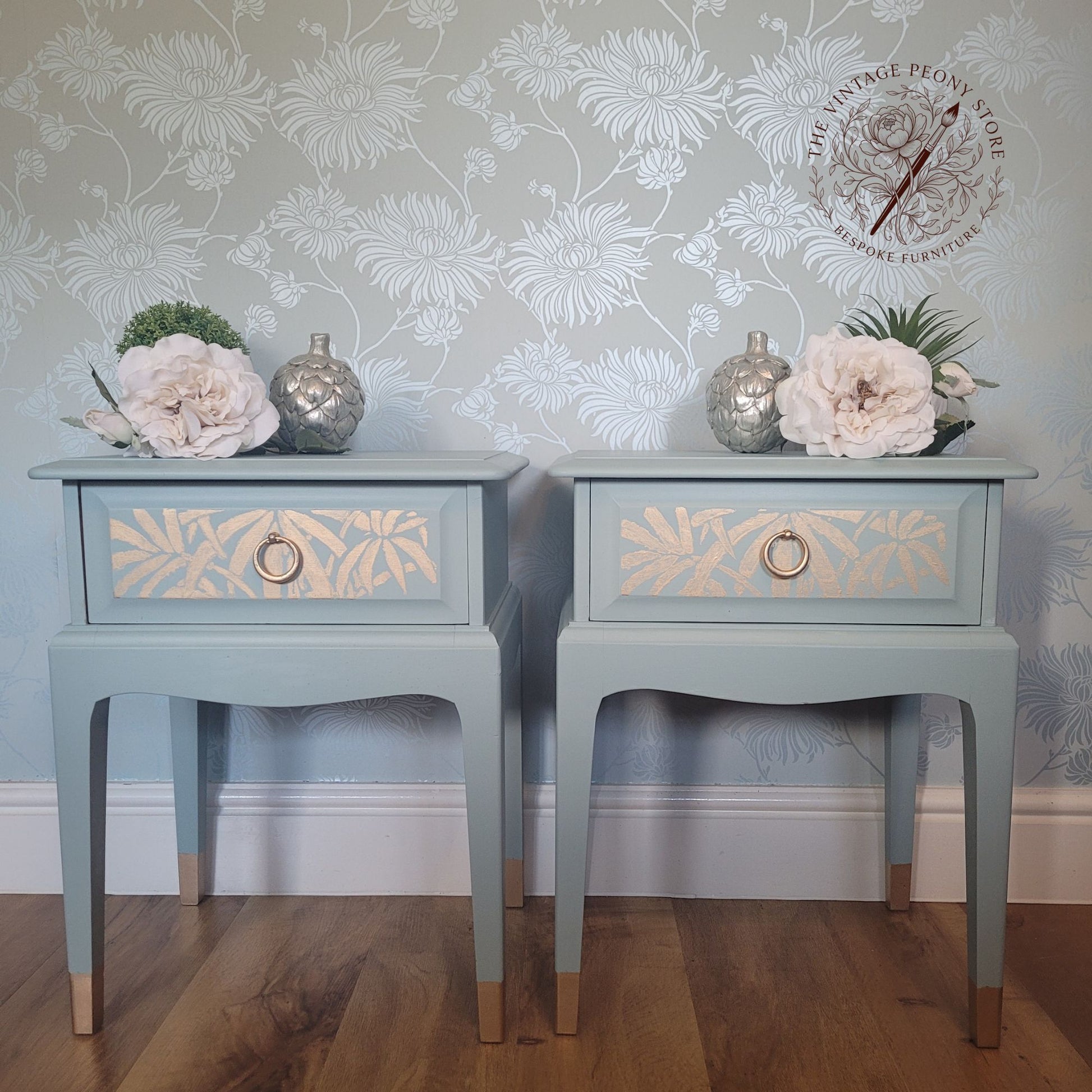 A pair of matching painted nightstands with floral stencil detailing on the drawer fronts, styled with faux flowers and silver vases against a floral wallpaper background. A logo for 'The Vintage People Shop' is visible in the top right corner.