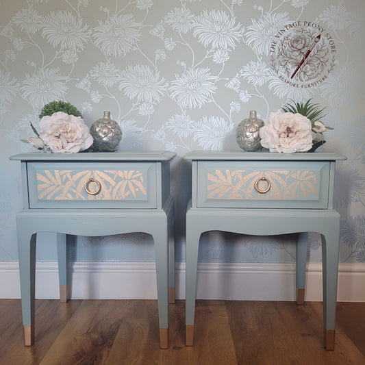A pair of matching painted nightstands with floral stencil detailing on the drawer fronts, styled with faux flowers and silver vases against a floral wallpaper background. A logo for 'The Vintage People Shop' is visible in the top right corner.