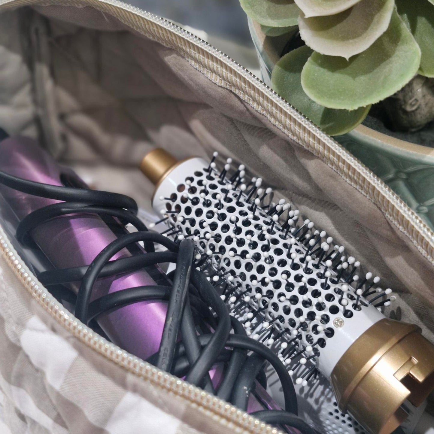 Hair dryer and round brush in a sand coloured gingham hair tool bag with a plant in the background