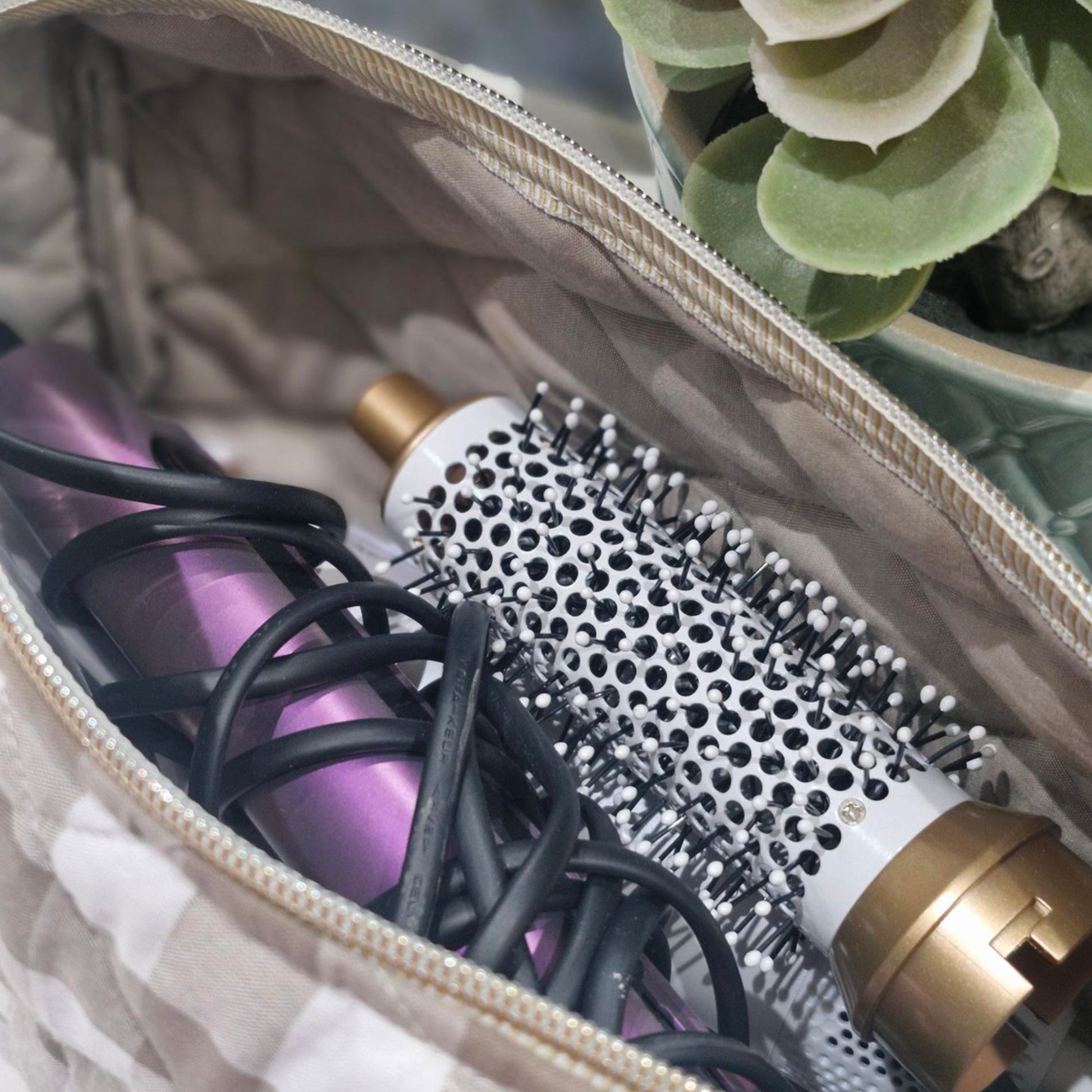 Hair dryer and round brush in a sand coloured gingham hair tool bag with a plant in the background