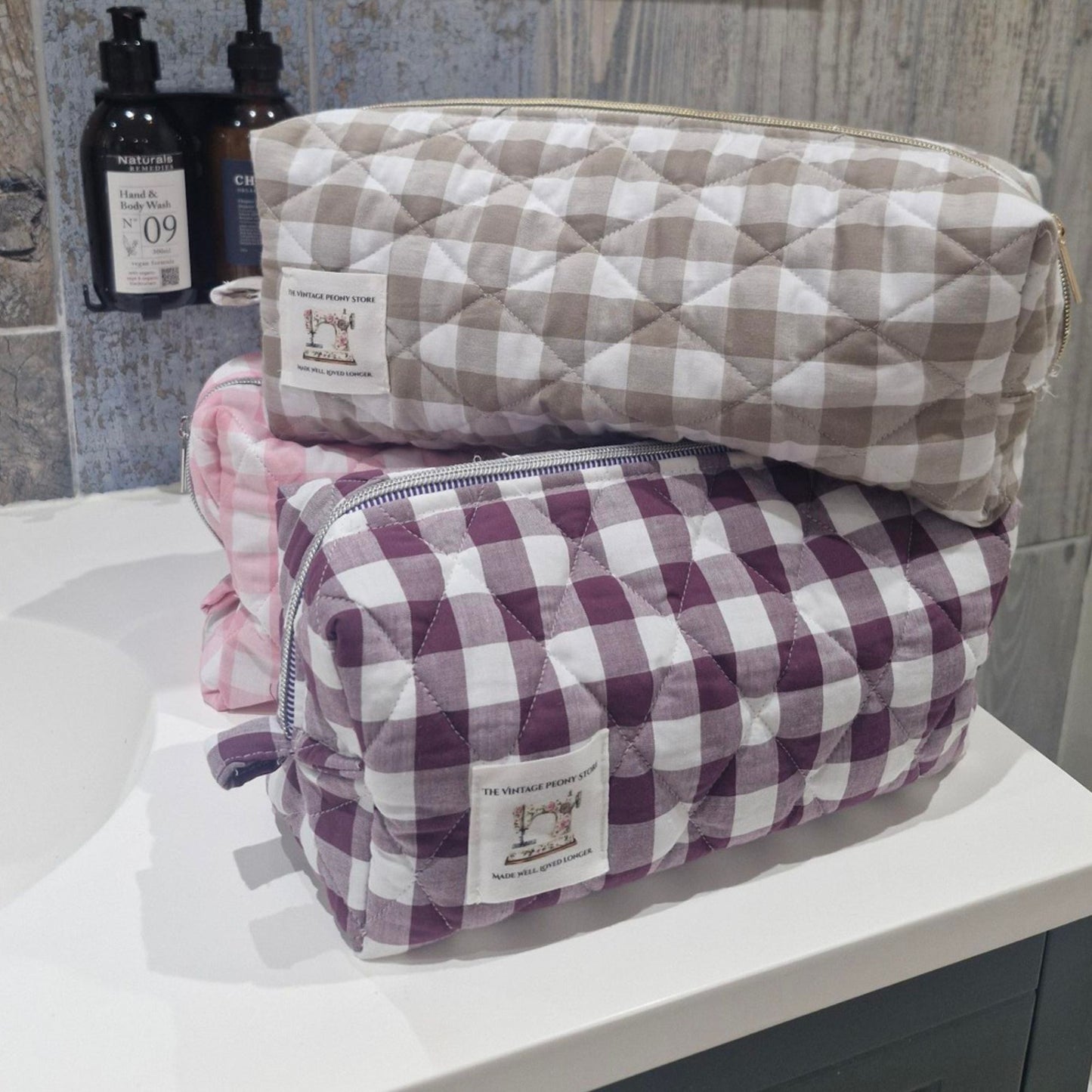 Three quilted hair tool bags, one in sand, one plum and one in a candy pink gingham colourway, stacked on a bathroom vanity unit.