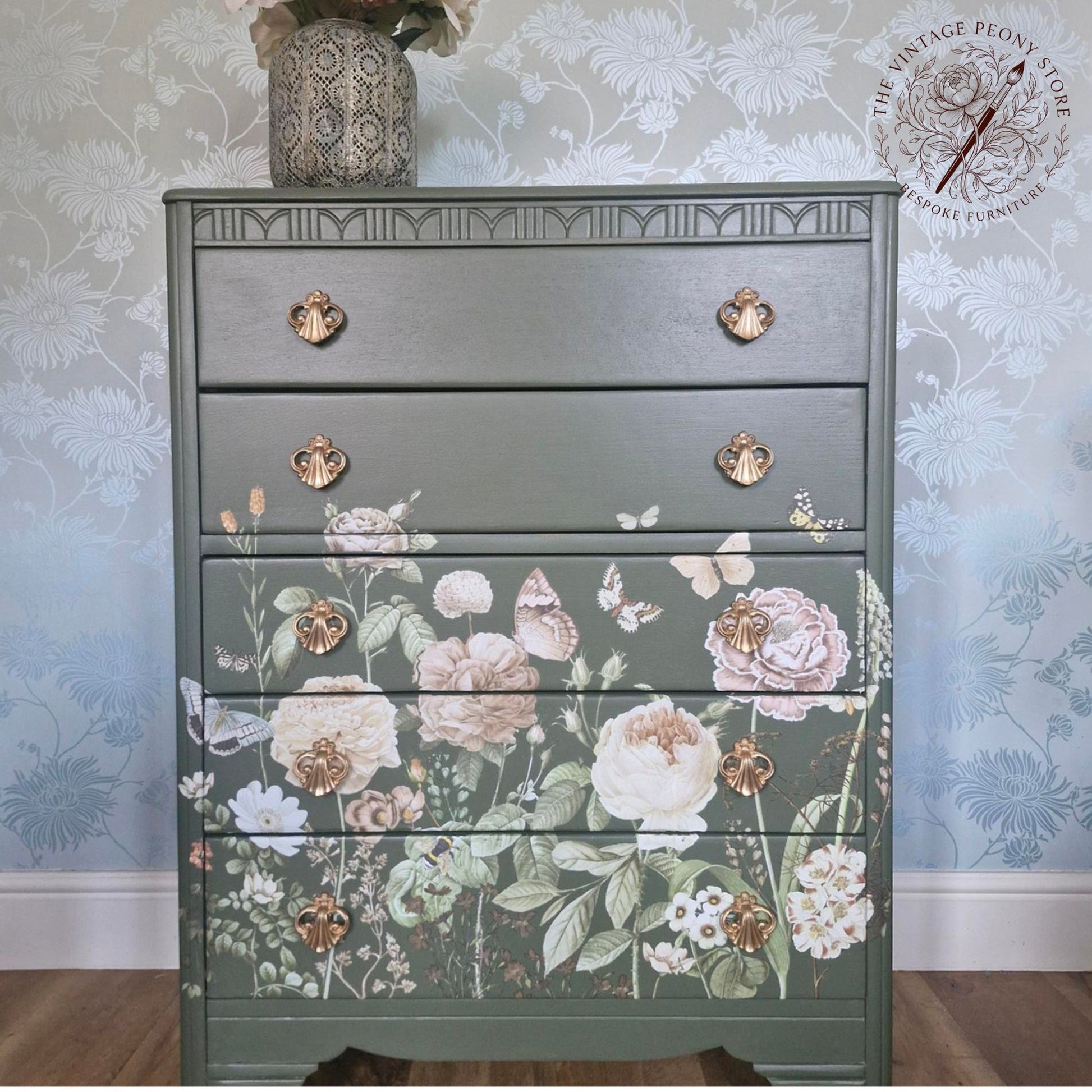 A painted five-drawer dresser in a muted sage green with a floral and butterfly design on the lower drawers, styled in a room with floral wallpaper and wooden flooring.