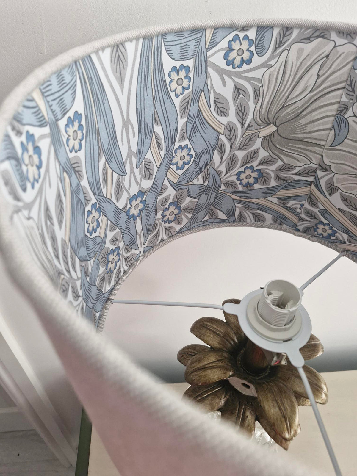 A close-up view inside a lampshade showing a blue, gray, and white floral pattern inspired by William Morris' style, with a brass-colored metal flower-shaped lamp base visible at the bottom.