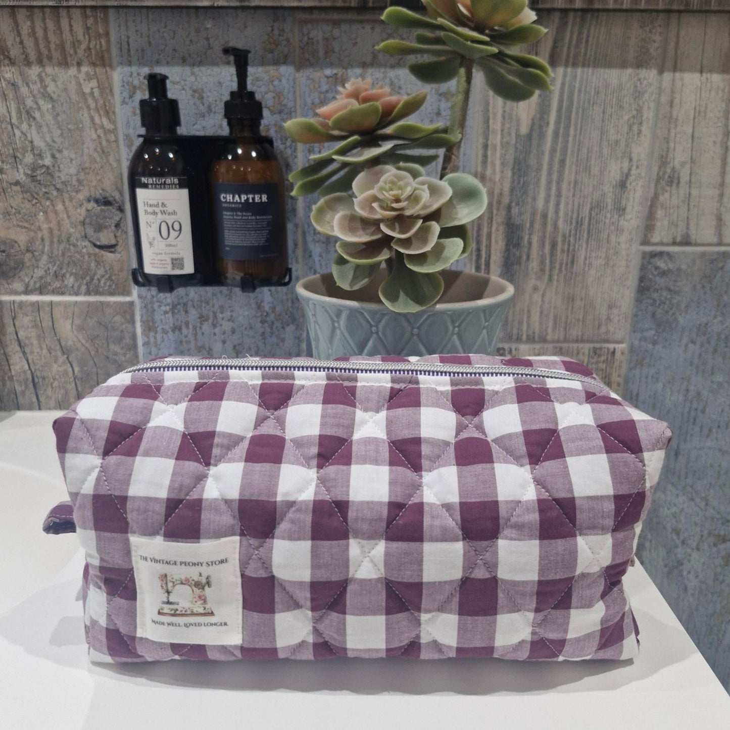 Quilted toiletry bag with checkered pattern on a surface with bottles and a plant in the background.
