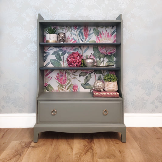 Beautifully upcycled green bookshelf with floral wallpaper backing. The shelves display decorative items and the bottom drawer features elegant gold ring pulls. Perfect for adding a touch of nature-inspired charm to any living room, bedroom, or hallway.