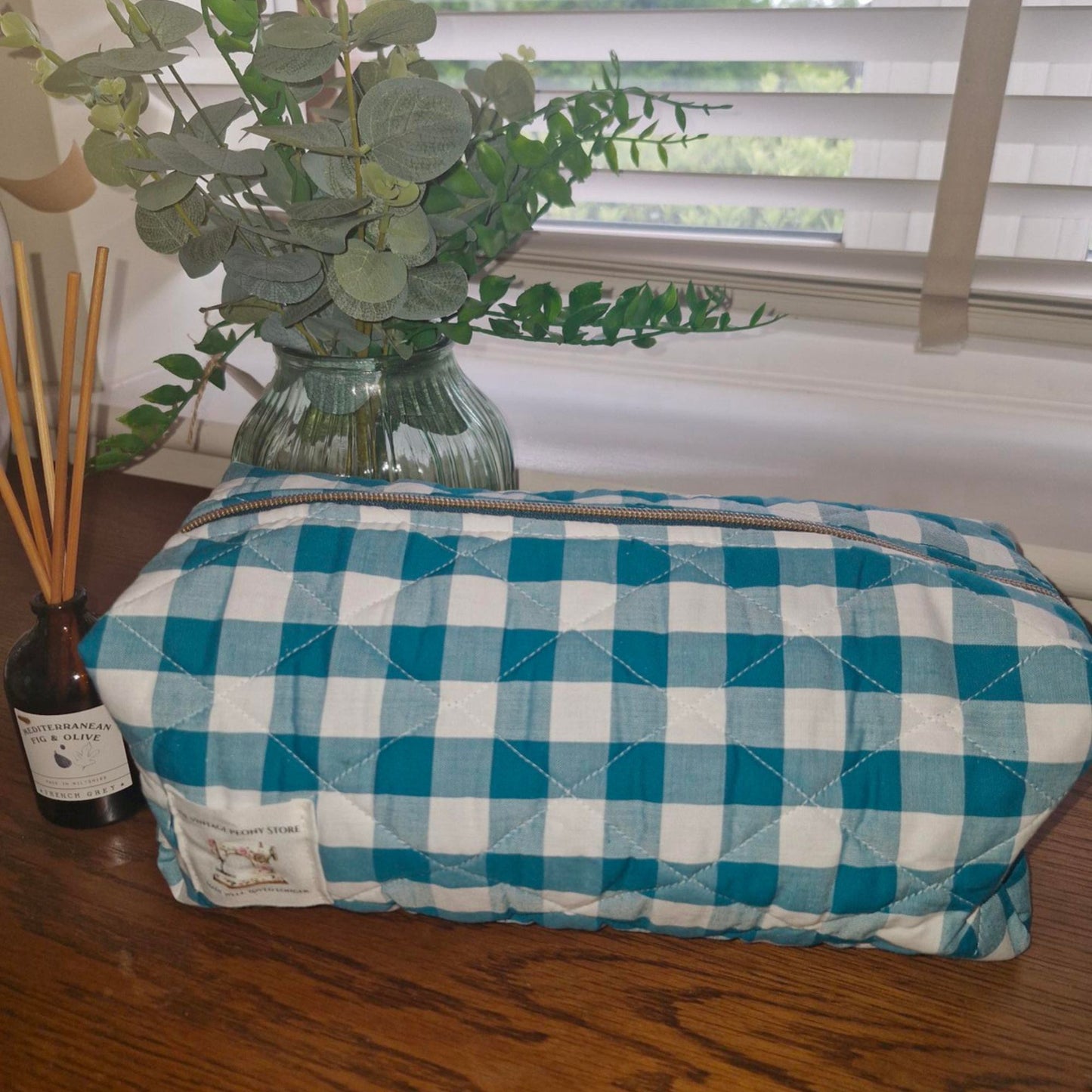 Teal quilted gingham hair tool bag on a surface with a vase of greenery and a diffuser in the background.