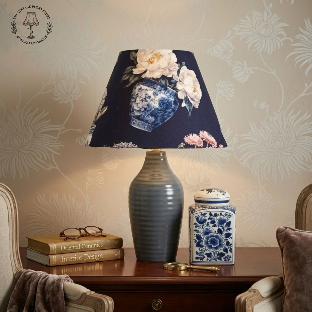 A table lamp with a dark navy blue fabric shade featuring a blue and white porcelain vase and floral print, paired with a ribbed gray ceramic base, set on a wooden table in a styled room.