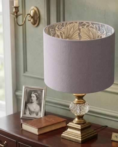 A table lamp with a lavender-gray fabric drum shade, a gold-toned metal base, and a clear faceted crystal pedestal, shown on a dark wooden table in a styled interior setting.
