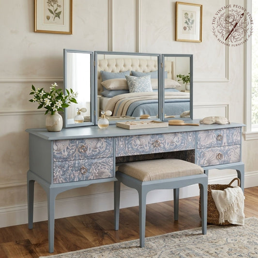 A vintage-style vanity table with a matching stool, featuring a light blue-gray painted frame and ornate floral-patterned drawer fronts. The vanity is styled with a three-panel mirror, a vase of white flowers, and a small mirror, set in a beautifully decorated bedroom.